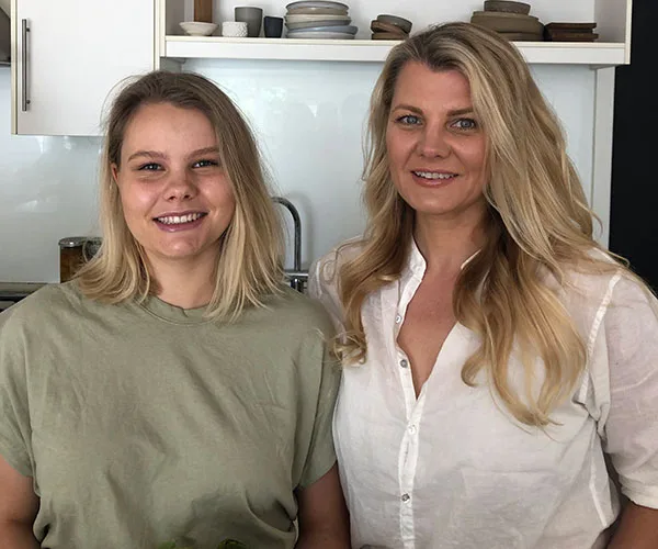 Two women smiling in a kitchen, one in a green shirt and the other in a white blouse, with shelves in the background.