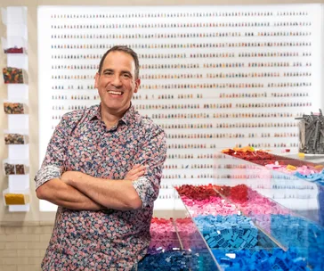 Man in a floral shirt smiling, standing in front of a wall display of LEGO minifigures, with colorful LEGO pieces in front.