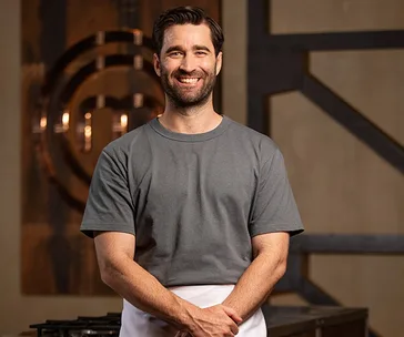 Man in a gray t-shirt and white apron smiling on a cooking show set.