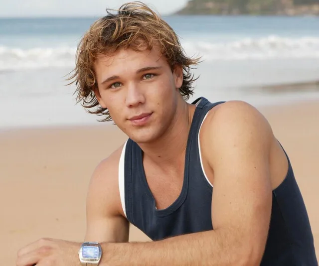Young man with short hair at the beach, wearing a navy tank top and a watch, with waves in the background.