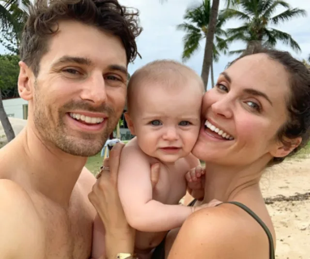 A couple smiling on a tropical beach, holding a baby, with palm trees in the background.