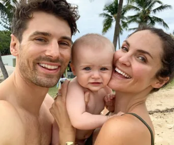 A couple smiling on a tropical beach, holding a baby, with palm trees in the background.