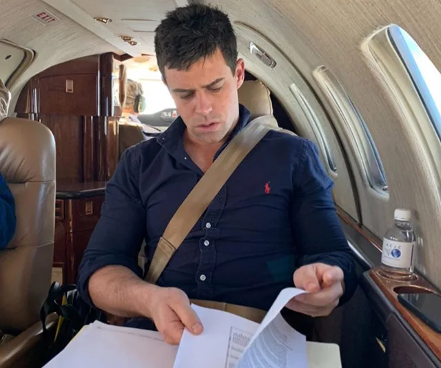 Man in a private jet reading documents, wearing a seatbelt and a dark shirt, with bottled water nearby.
