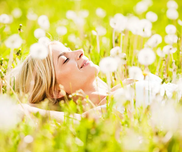 Woman lying in a field of dandelions, smiling with eyes closed, enjoying the sunshine.