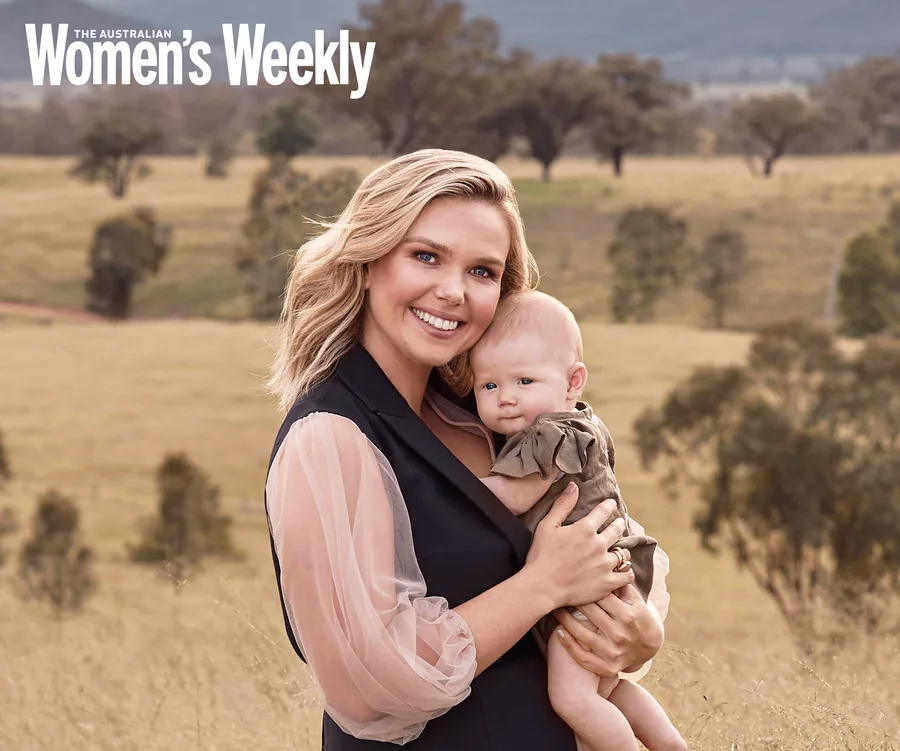 "Smiling woman holding baby in a field, featured in The Australian Women's Weekly magazine."