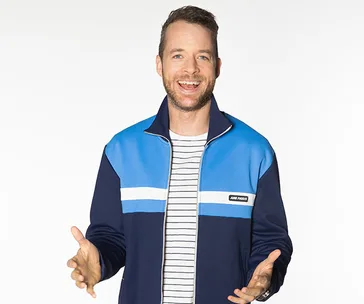 A man in a blue and black jacket with a striped shirt, smiling and gesturing against a plain white background.