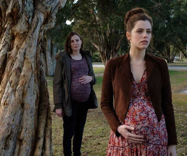 Two pregnant women stand in a park, one in brown jacket and floral dress, the other in a green coat.