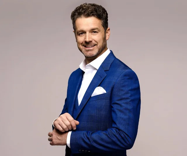 A man in a blue suit with a white shirt and pocket square, smiling, standing against a plain background.
