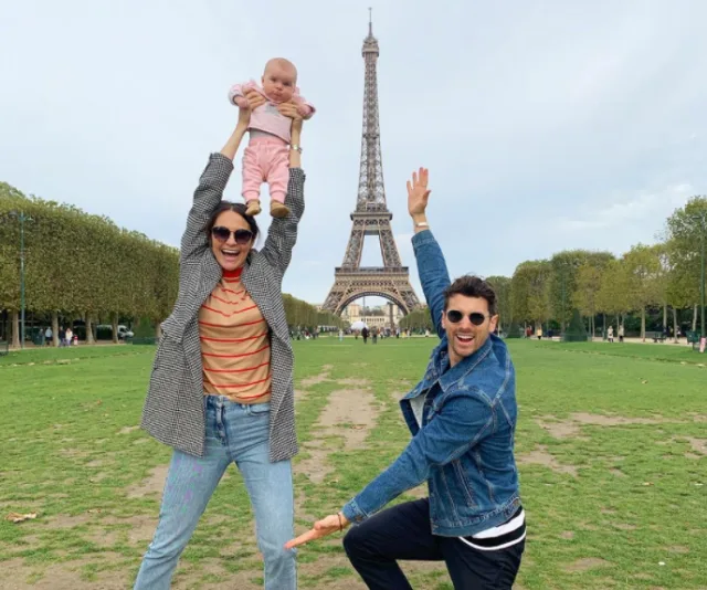 A couple with a baby in front of the Eiffel Tower, smiling and posing playfully on the green lawn.