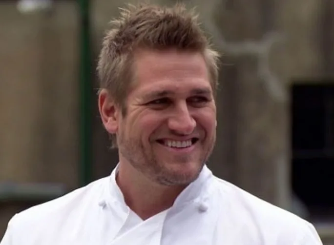 Celebrity chef in white uniform smiling, outdoors.