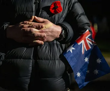 Hands clasped, red poppy on jacket, holding Australian flag, honoring ANZAC Day.