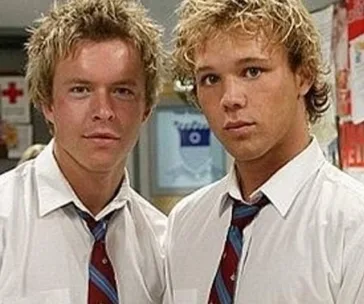 Two young men with blond hair in school uniforms with ties, standing together in a school hallway.