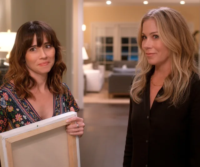 Two women from "Dead to Me" stand in a warmly lit room; one holds a canvas, both look intrigued.