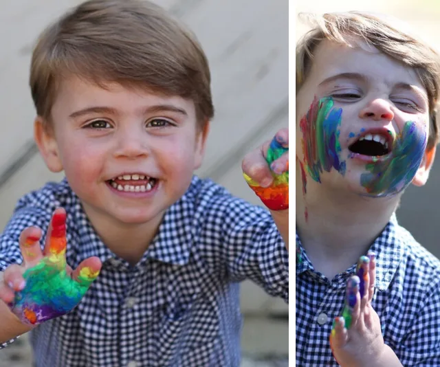 A young child with a checkered shirt joyfully shows painted hands and face, smiling in an outdoor setting.