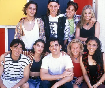 A group photo of eight smiling young adults from the show "Heartbreak High," posing together outdoors.