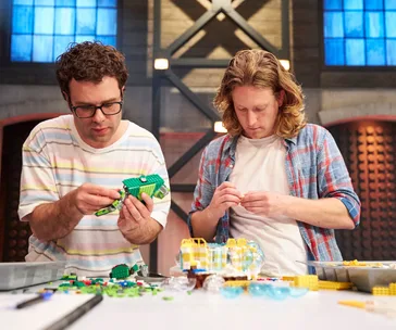 Two individuals focused on building LEGO creations, surrounded by colored bricks on a table in a studio setting.