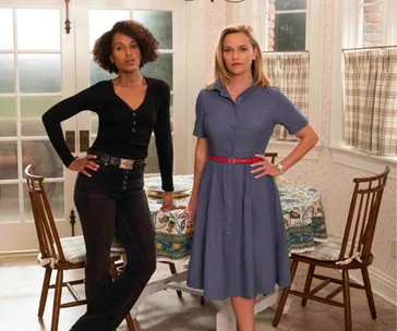 Two women stand confidently in a kitchen, one in a black outfit, the other in a blue dress.