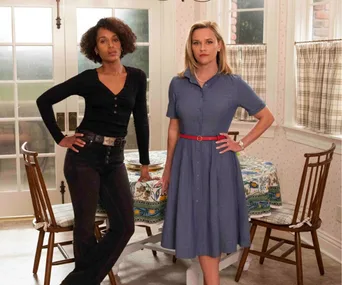 Two women stand confidently in a kitchen, one in a black outfit, the other in a blue dress.