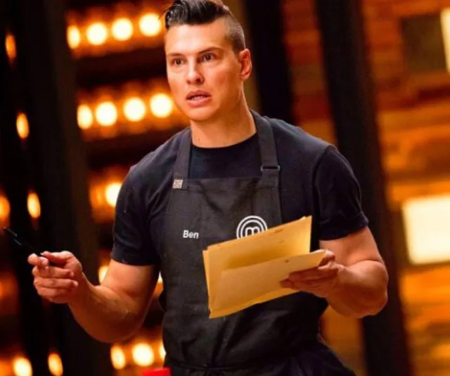 Chef in black apron holding a notebook and pen, focused expression, bright kitchen background.