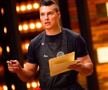 Chef in black apron holding a notebook and pen, focused expression, bright kitchen background.