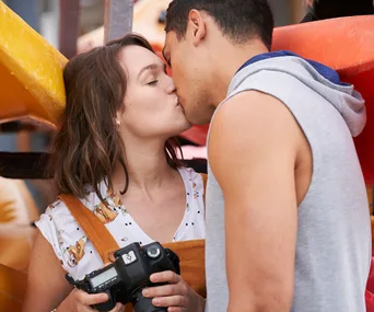 A couple shares a kiss; the woman holds a camera, wearing patterned overalls. Kayaks are stacked in the background.
