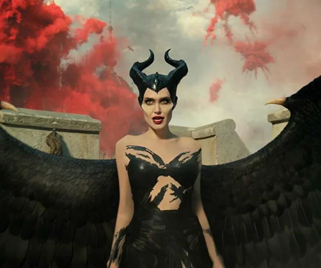 Maleficent with dark wings and horned headpiece stands against a cloudy sky with red smoke in the background.