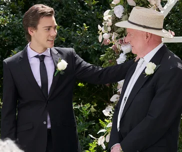 Two men in suits, one with a hat, smiling at each other at an outdoor event with flowers in the background.