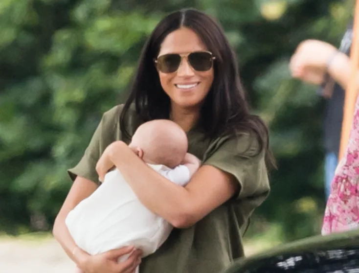 Woman in sunglasses smiling while holding a baby outdoors.