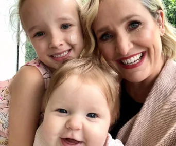 A woman smiling with two young children, a toddler and a baby, all appearing happy together.