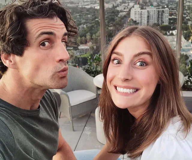 Man and woman smiling on a balcony with cityscape in the background.