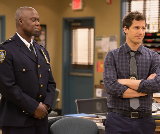 Two men, one in a police uniform and another in casual office attire, stand in a police precinct setting.