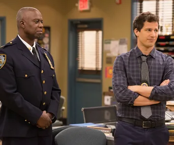 Two men, one in a police uniform and another in casual office attire, stand in a police precinct setting.