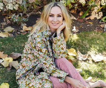 Blonde woman in floral dress sits on grass, smiling with hand on chin, surrounded by autumn leaves and flowers.
