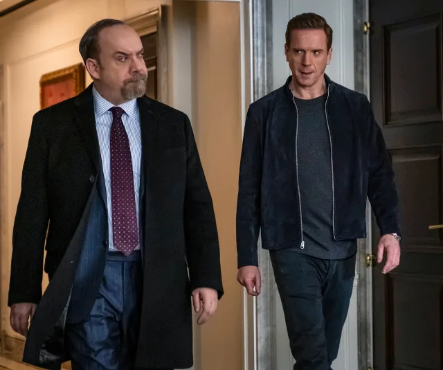Two men walking in a hallway, one in a suit, the other in casual wear, from a scene in the show "Billions."