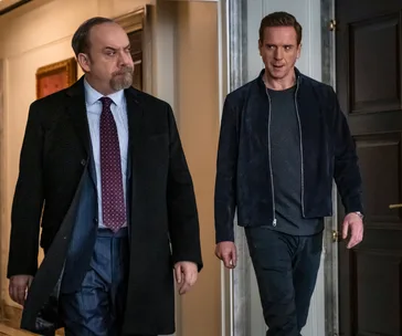 Two men walking in a hallway, one in a suit, the other in casual wear, from a scene in the show "Billions."