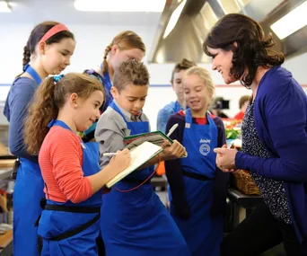 Children in blue aprons working with an adult in a kitchen, likely participating in a cooking activity or competition.