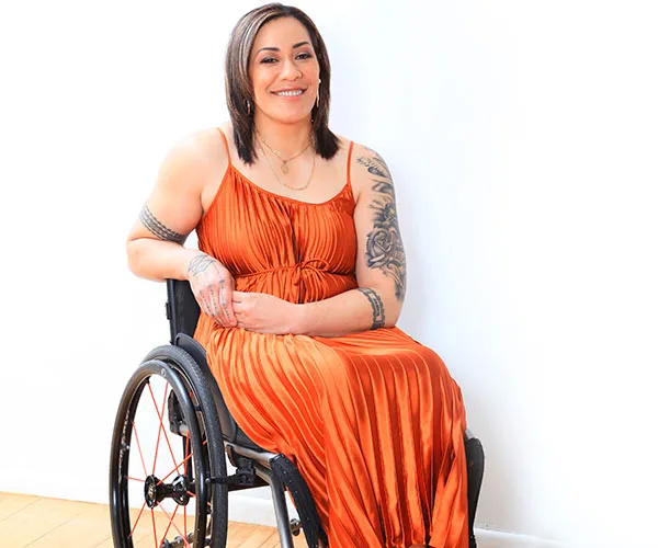 A woman in an orange dress sits in a wheelchair, smiling, with tattoos on her arms.