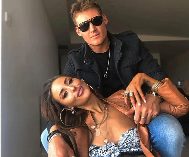 A couple poses together, the man wearing sunglasses, while the woman leans on him, both smiling.