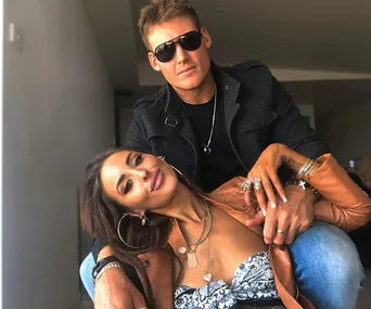 A couple poses together, the man wearing sunglasses, while the woman leans on him, both smiling.