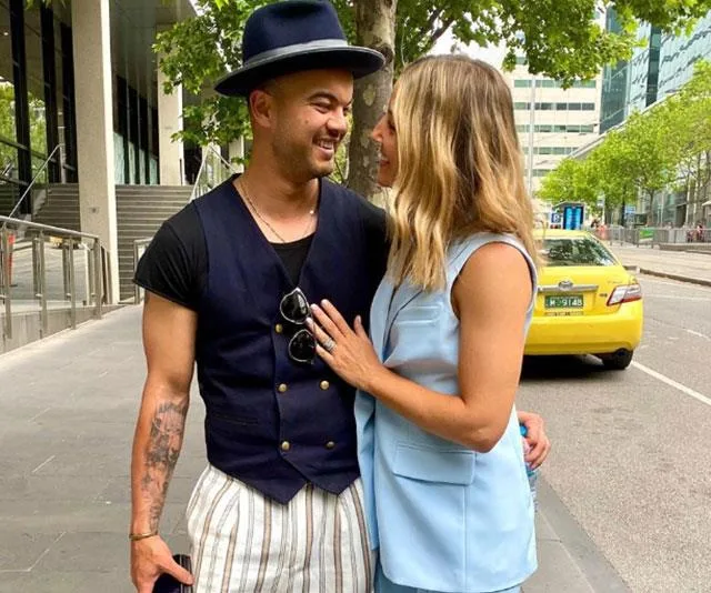 A couple smiling at each other on a city sidewalk; the man wears a hat and vest, and the woman is in a light blue outfit.