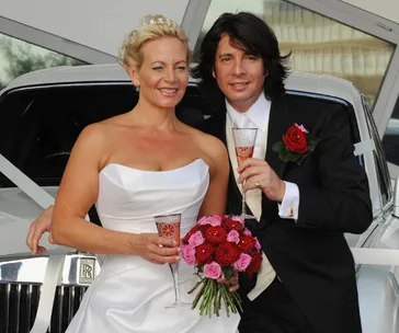 A couple in wedding attire celebrates with champagne by a car, holding a bouquet of red and pink roses.