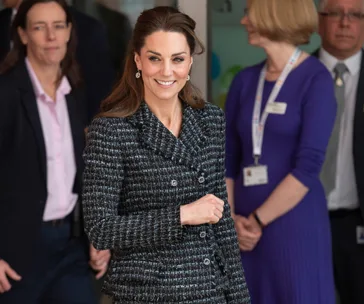 A woman in a checkered coat smiles while walking, with people standing in the background.