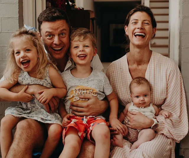 Happy family of four with young children and a baby, smiling and seated together at home.