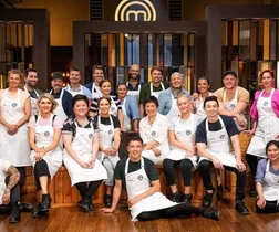 Group photo of "MasterChef All Stars" contestants in kitchen, wearing white aprons, posing together, and smiling.