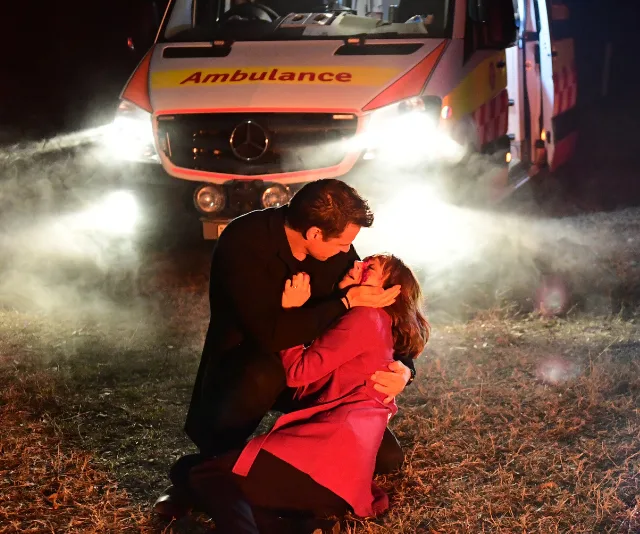 Man holds injured woman in his arms on a dark road, bright ambulance headlights shining in the background.