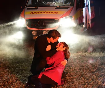 Man holds injured woman in his arms on a dark road, bright ambulance headlights shining in the background.