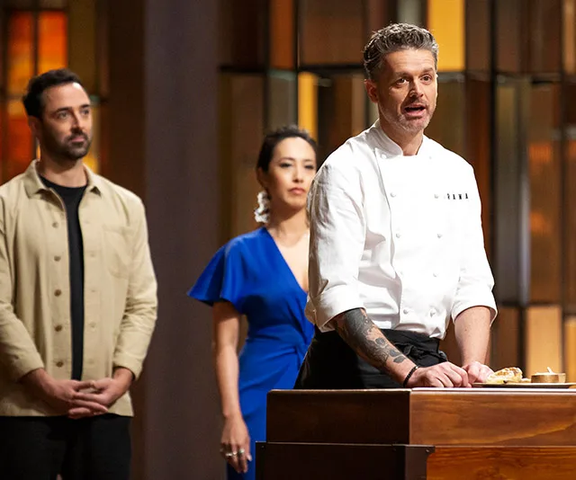 MasterChef judge speaking at podium, two people in the background.