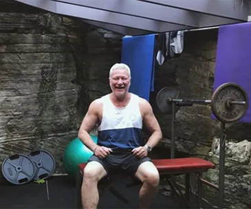 Man sitting on a bench in a gym setting, smiling, with weights in the background.