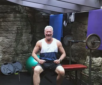 Man sitting on a bench in a gym setting, smiling, with weights in the background.