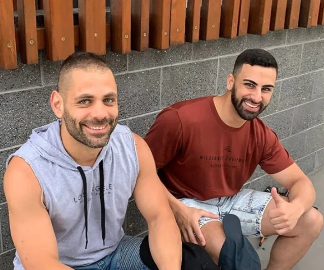 Two men sitting on the ground, smiling at the camera, against a wall with wooden panels. One is in a sleeveless hoodie.